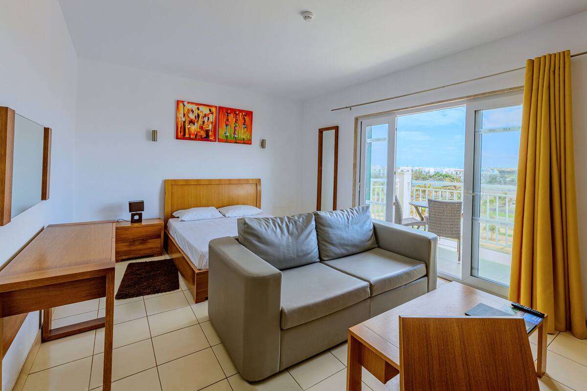 Une chambre d’hôtel moderne équipée d’un lit, d’un canapé, d’un bureau et d’un balcon avec des rideaux jaunes.