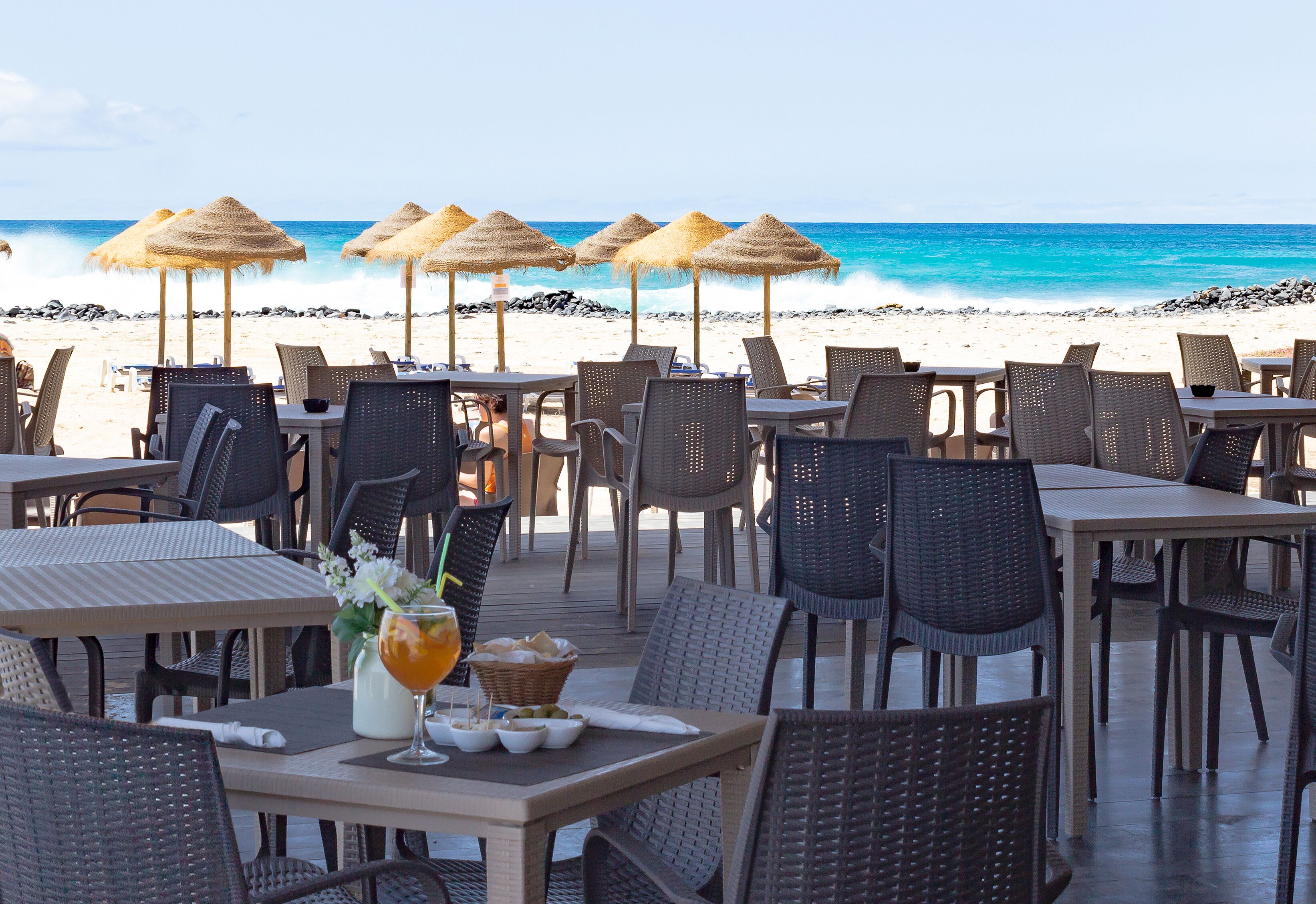 Un restaurante frente al mar con terraza, sombrillas de paja y vistas al océano.
