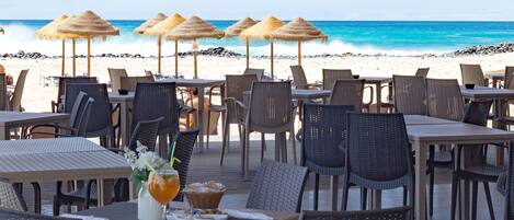 Un restaurante frente al mar con terraza, sombrillas de paja y vistas al océano.