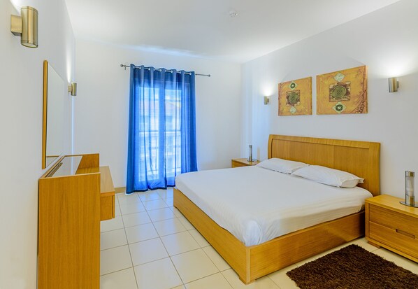 A bedroom with a wooden bed, a nightstand, a desk, and a large window with blue curtains.