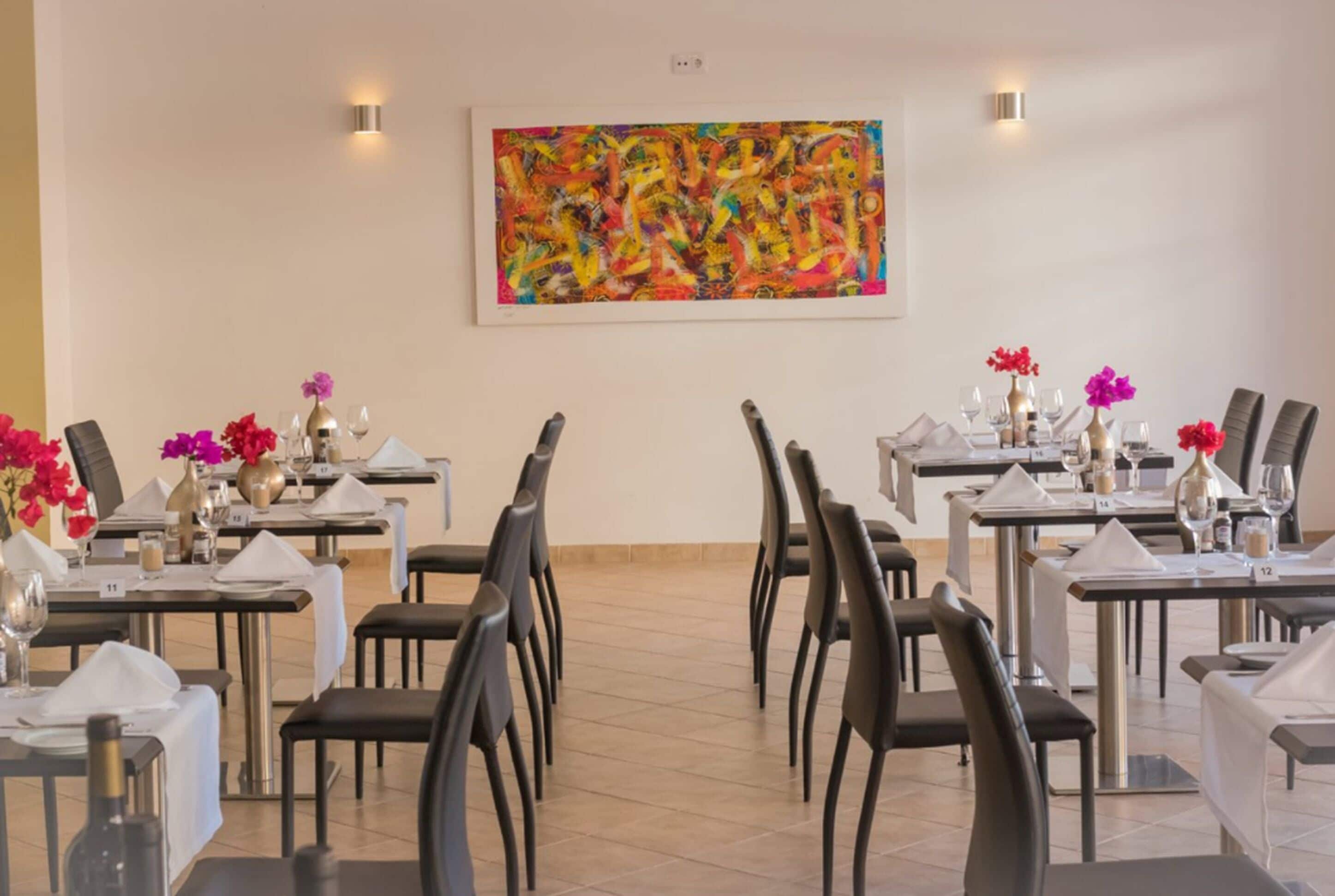 A dining area with tables set for service, a large abstract painting on the wall, and decorative flowers.