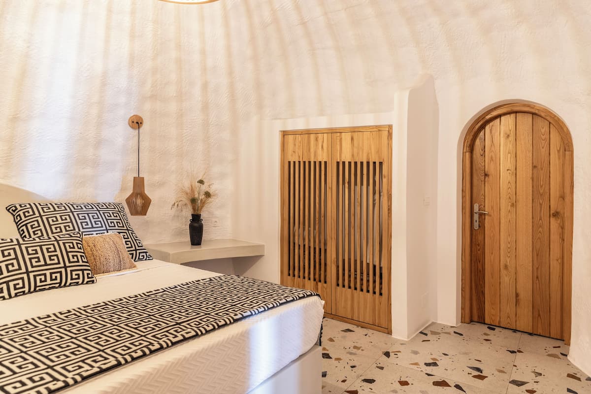 A bedroom with a bed, patterned pillows, a wooden door, and a wall-mounted light fixture.