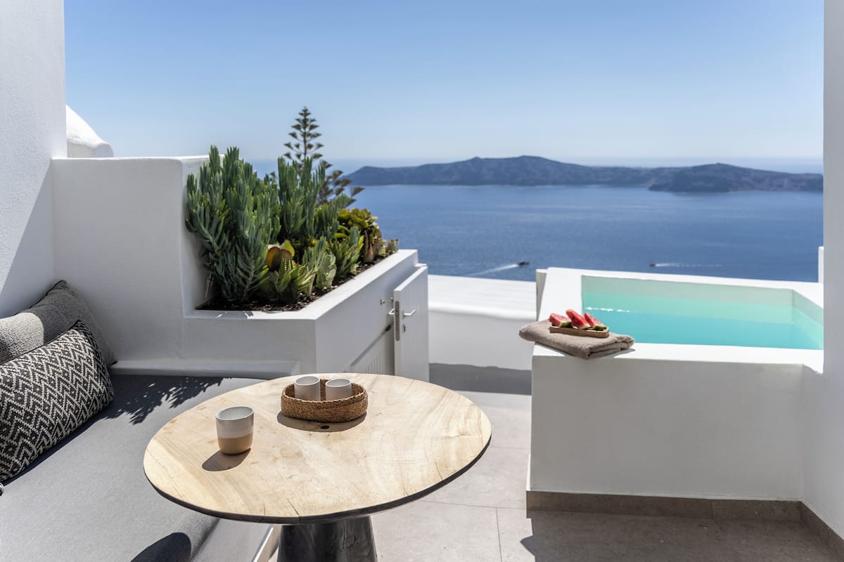 A balcony with a round table, a small seating area with a cushion, a plant arrangement, and a view of the sea.