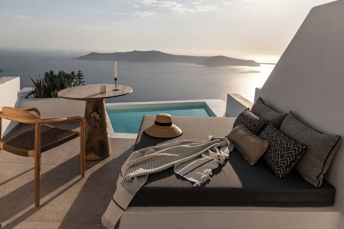 A sunlit terrace with a pool, a wooden chair, a round table, and a sofa with cushions.