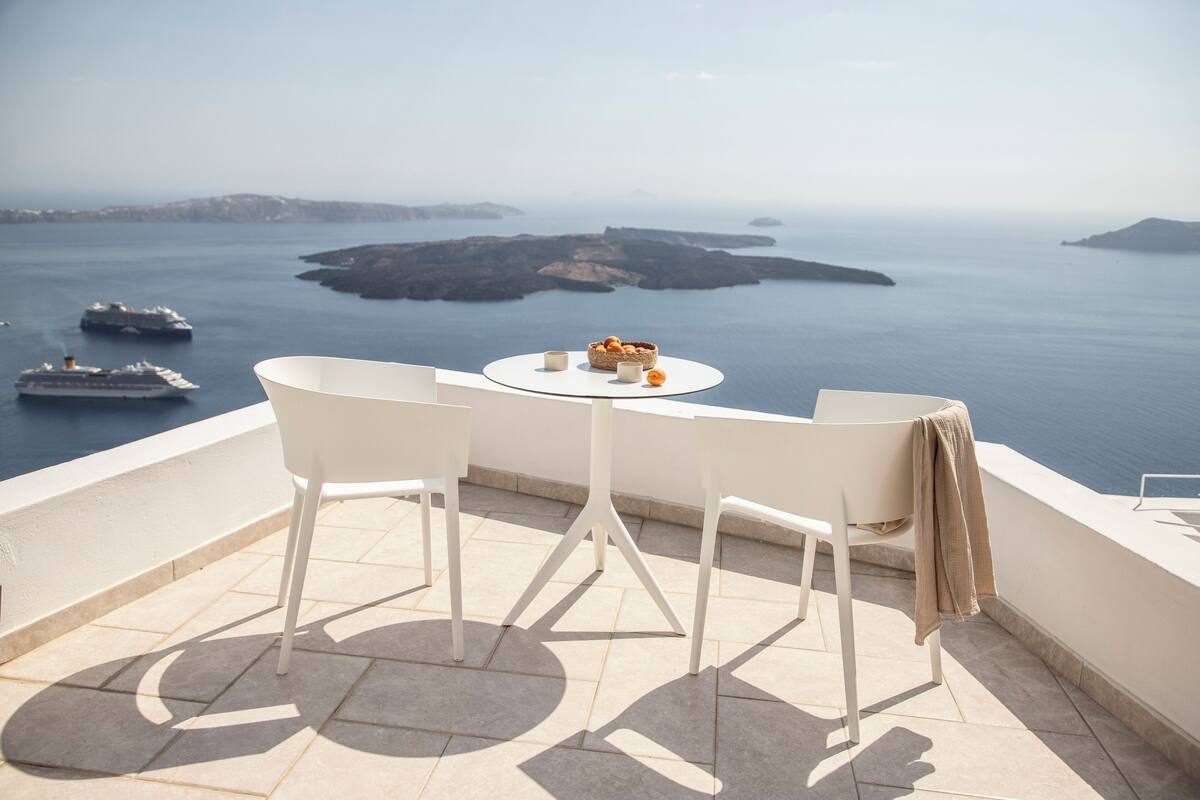 A terrace with two white chairs and a small table, overlooking a body of water with ships and islands.