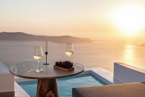 A round wooden table with two wine glasses and a candle, overlooking a pool and a sunset view. - LUKAS Aethereal Living (Santorini)