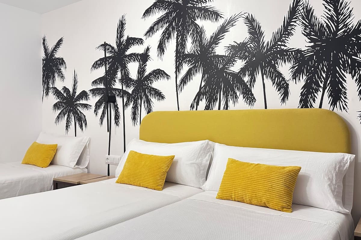 Habitación de hotel con dos camas, cada una con ropa blanca y almohadas amarillas, y un mural grande en la pared con palmeras.