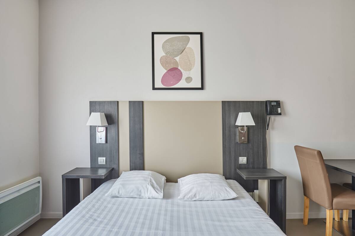 Quarto de hotel com uma cama, duas mesas de cabeceira com abajures, uma cadeira e uma obra de arte abstrata emoldurada na parede.