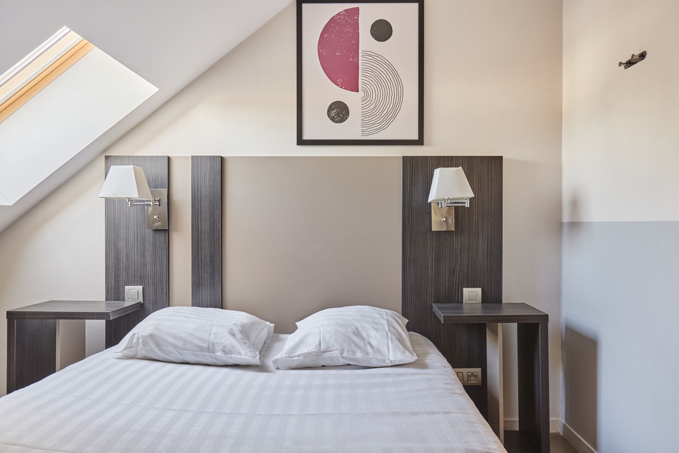 Een moderne hotelkamer met een groot bed, twee nachtkastjes met lampen, een dakraam en een abstract kunstwerk aan de muur.