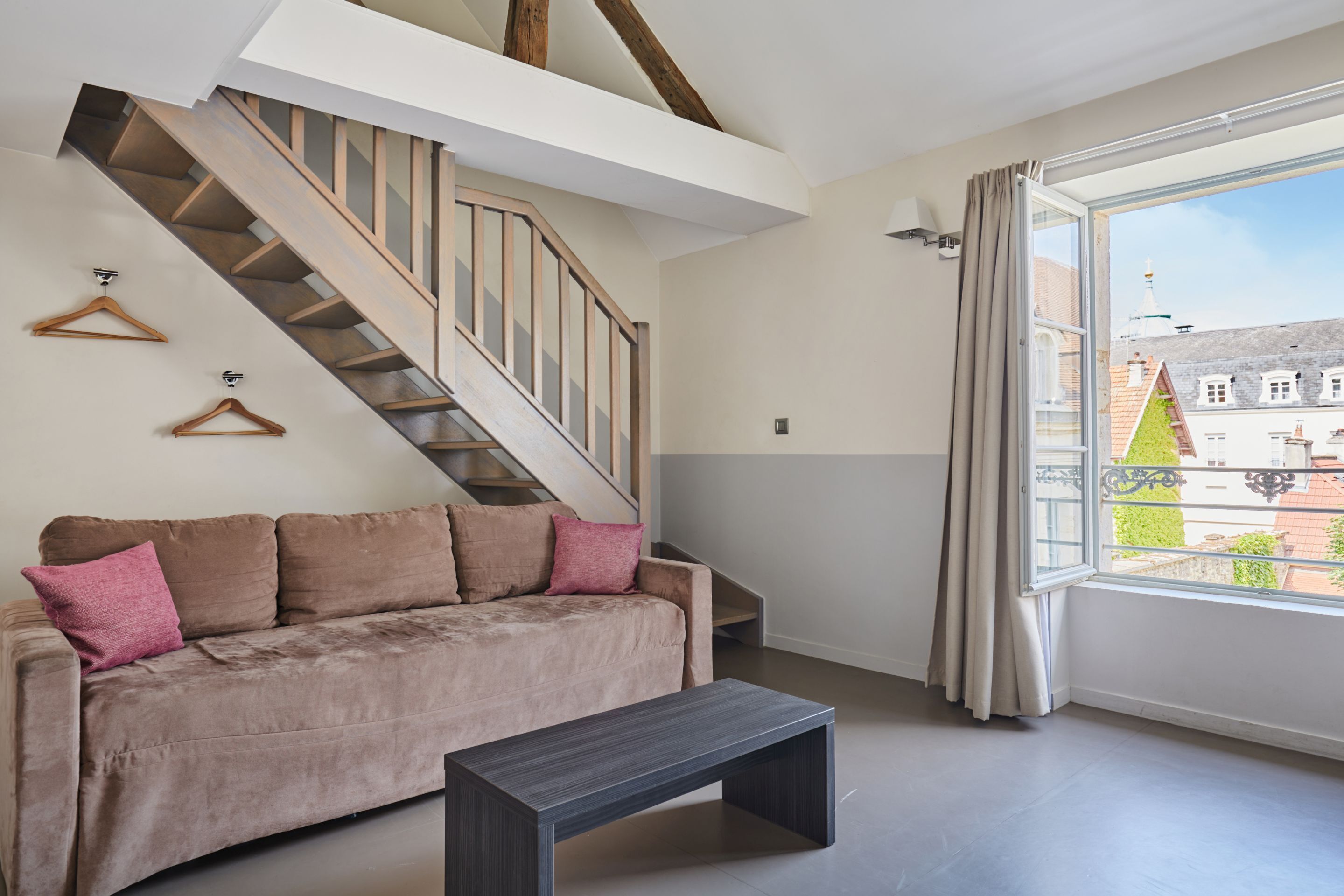 A modern living room with a staircase, a sofa, and a window with a view of buildings.