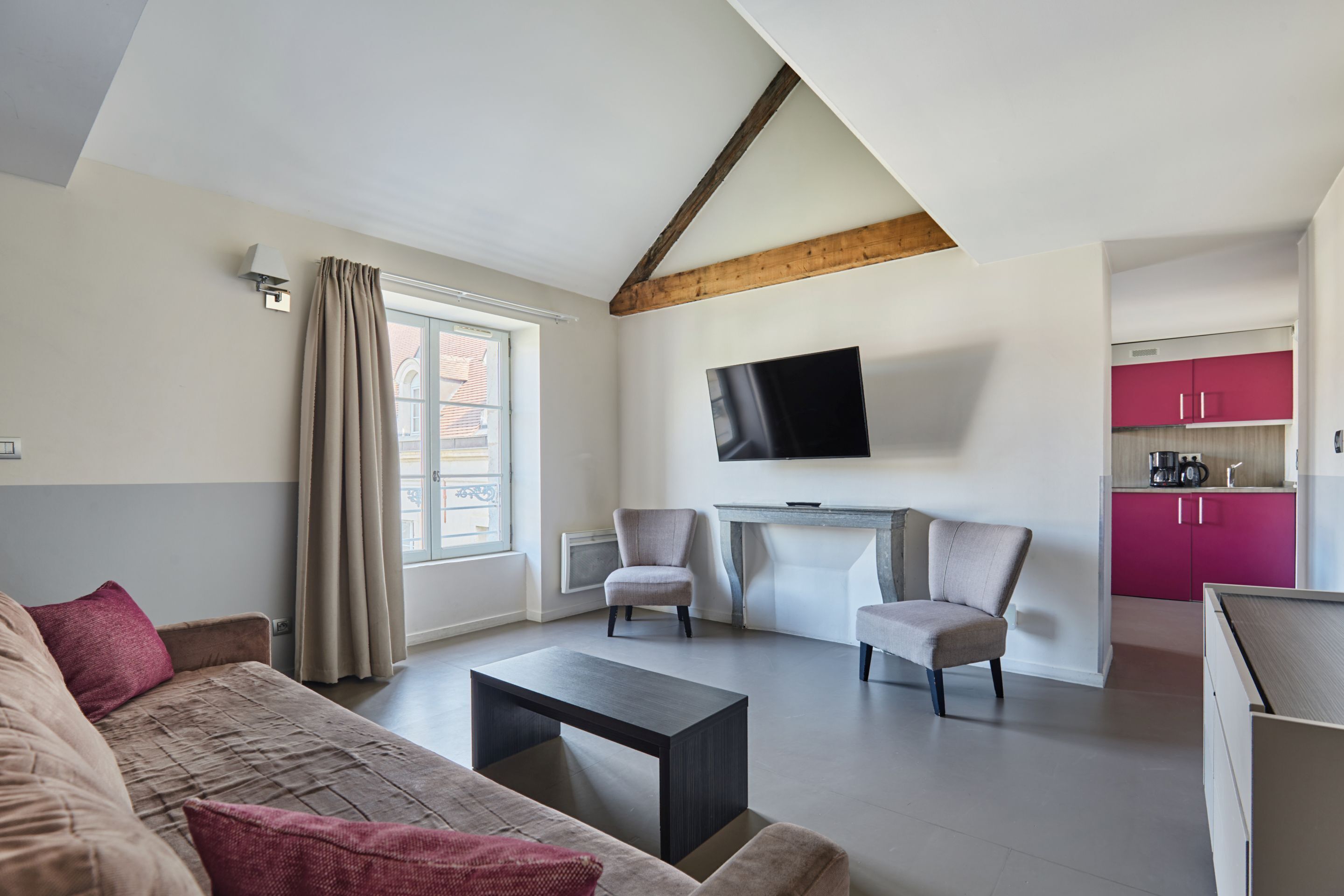 Una sala de estar moderna con un sofá, dos sillones, una mesa de centro y un televisor montado en la pared. Se observa un área de cocina con gabinetes rosas al fondo.