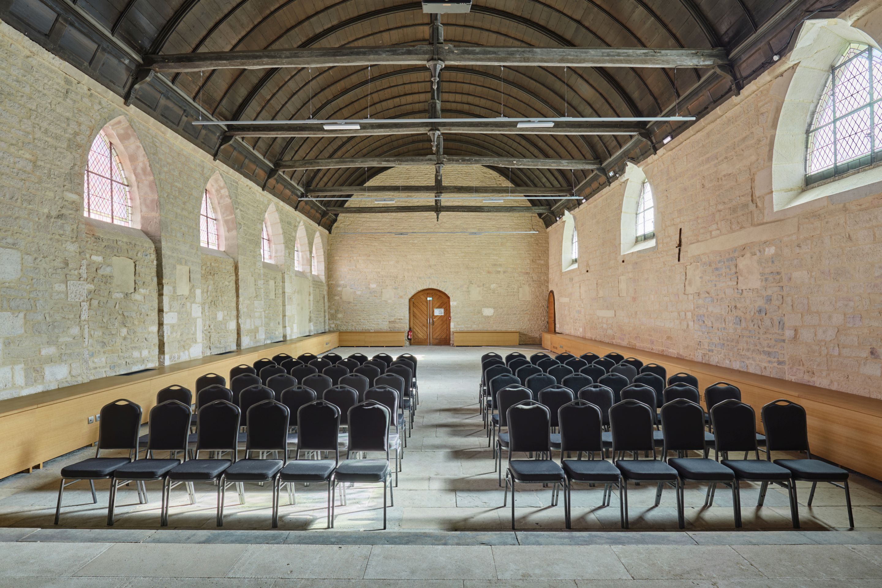 A large hall with rows of chairs facing a central door, stone walls, and arched windows.