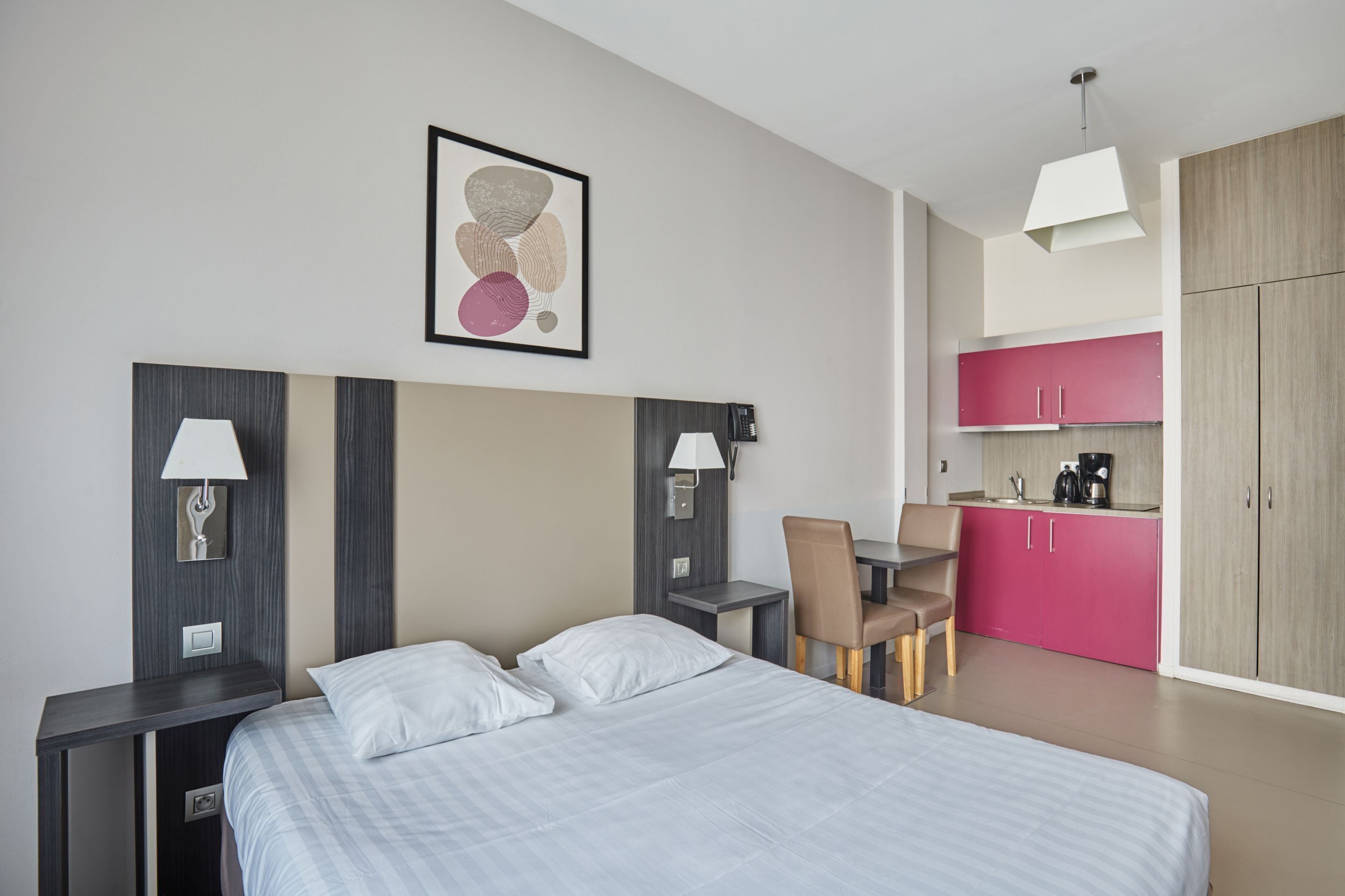 A modern hotel room with a large bed, bedside lamps, a desk, a chair, and a kitchenette with pink cabinets.