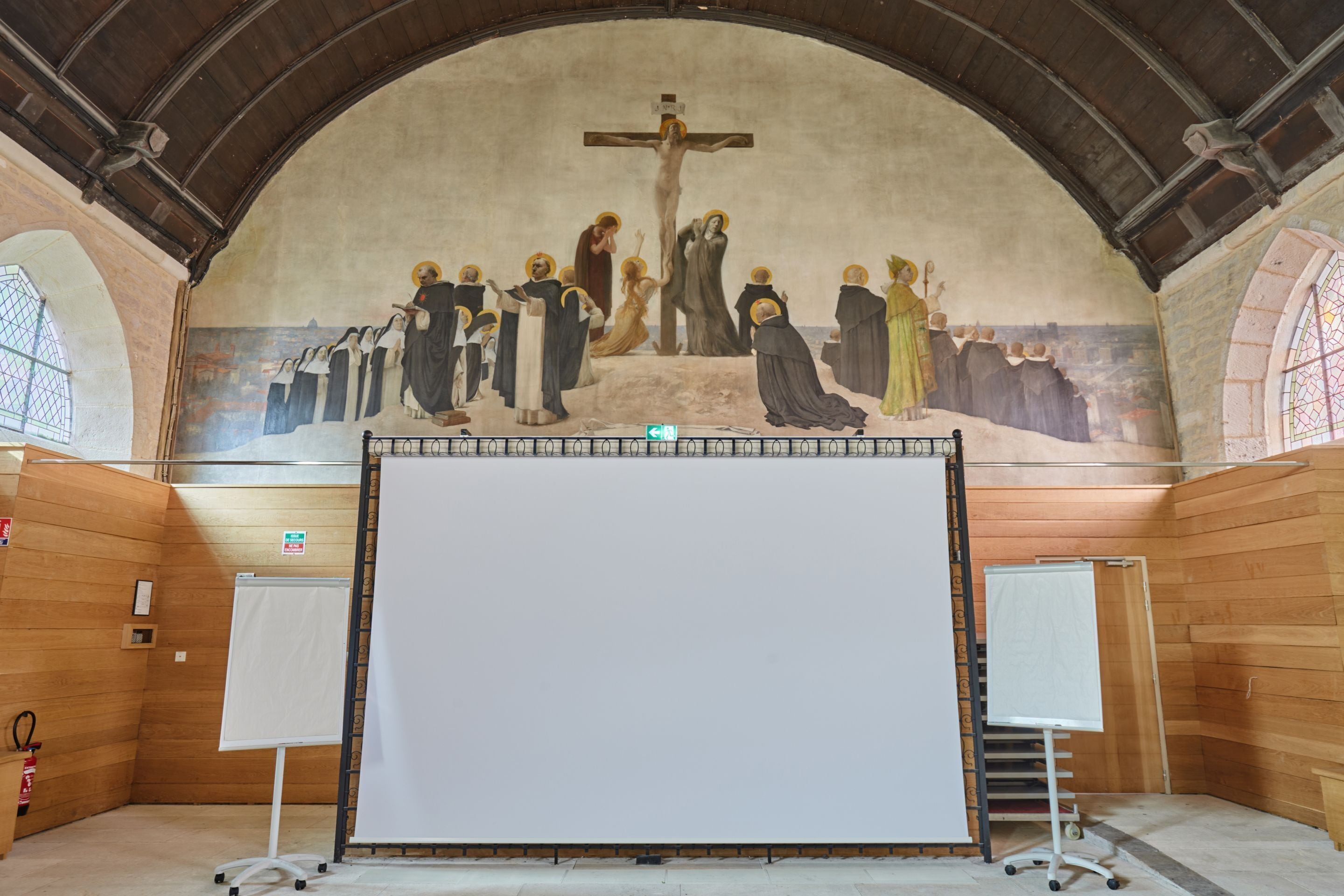 An interior with a large mural depicting a religious scene, wooden paneling, and a projection screen in the center.