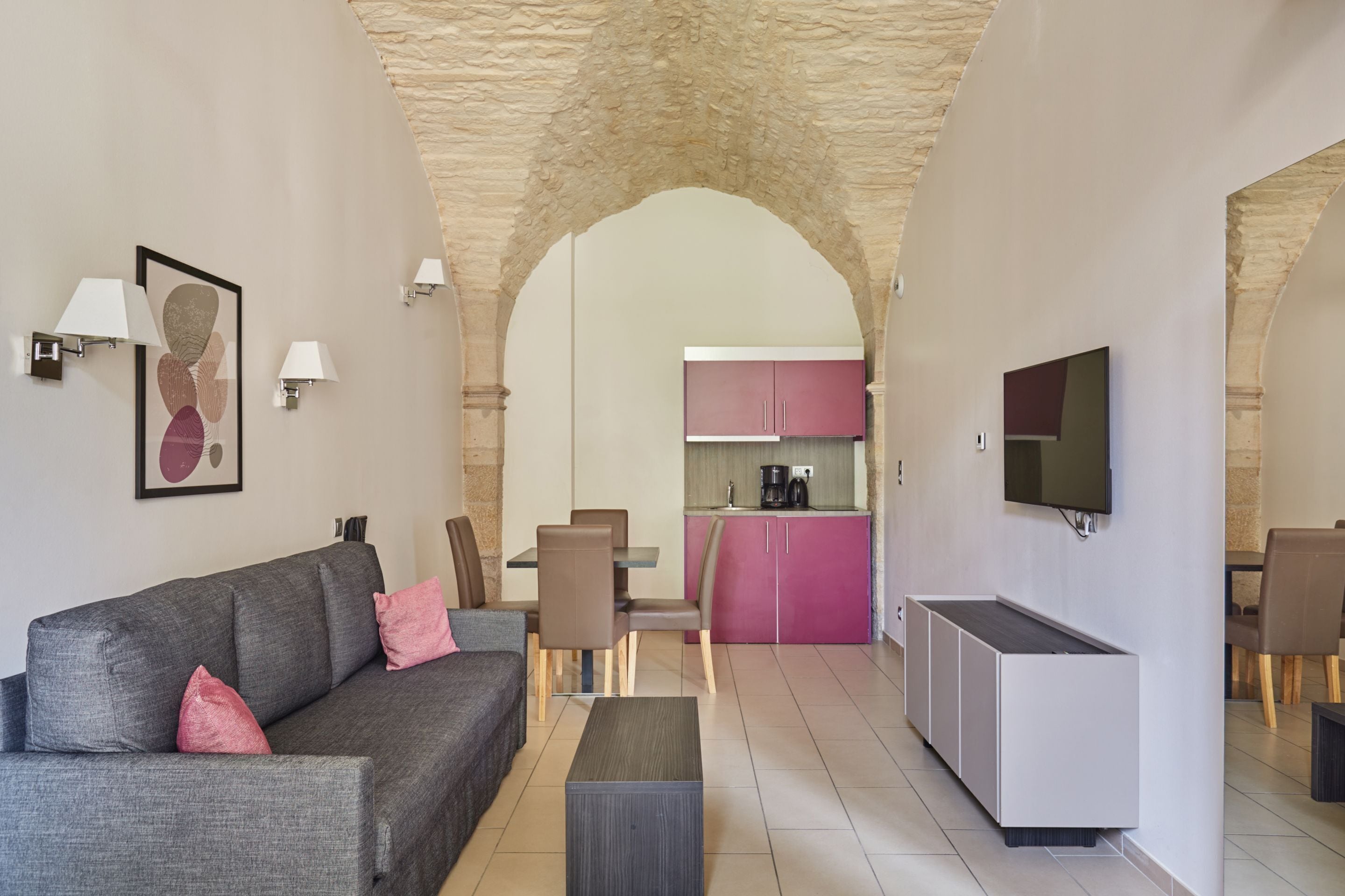 Ein modernes Wohnzimmer mit einem grauen Sofa, rosafarbenen Kissen, einem Couchtisch und einem an der Wand befestigten Fernseher.