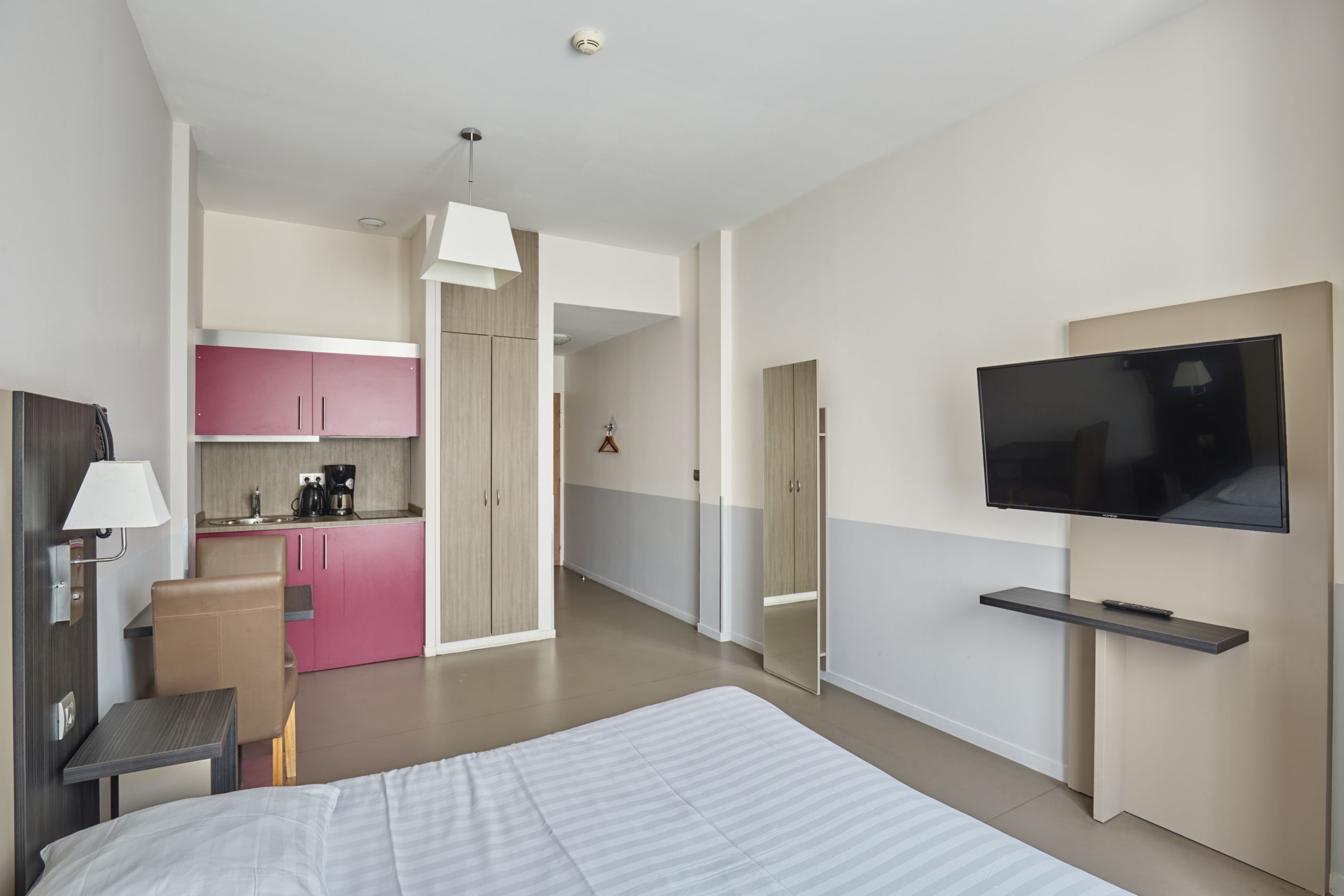 A modern hotel room with a bed, a desk, a chair, a TV, and a kitchenette.