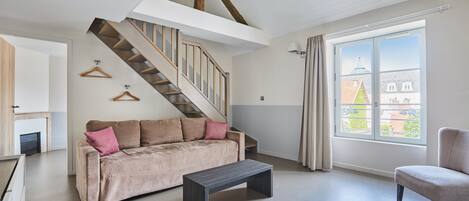 En rummelig stue med sofa, sofabord og lænestol. En trappe med træbjælker fører til en etage ovenover.