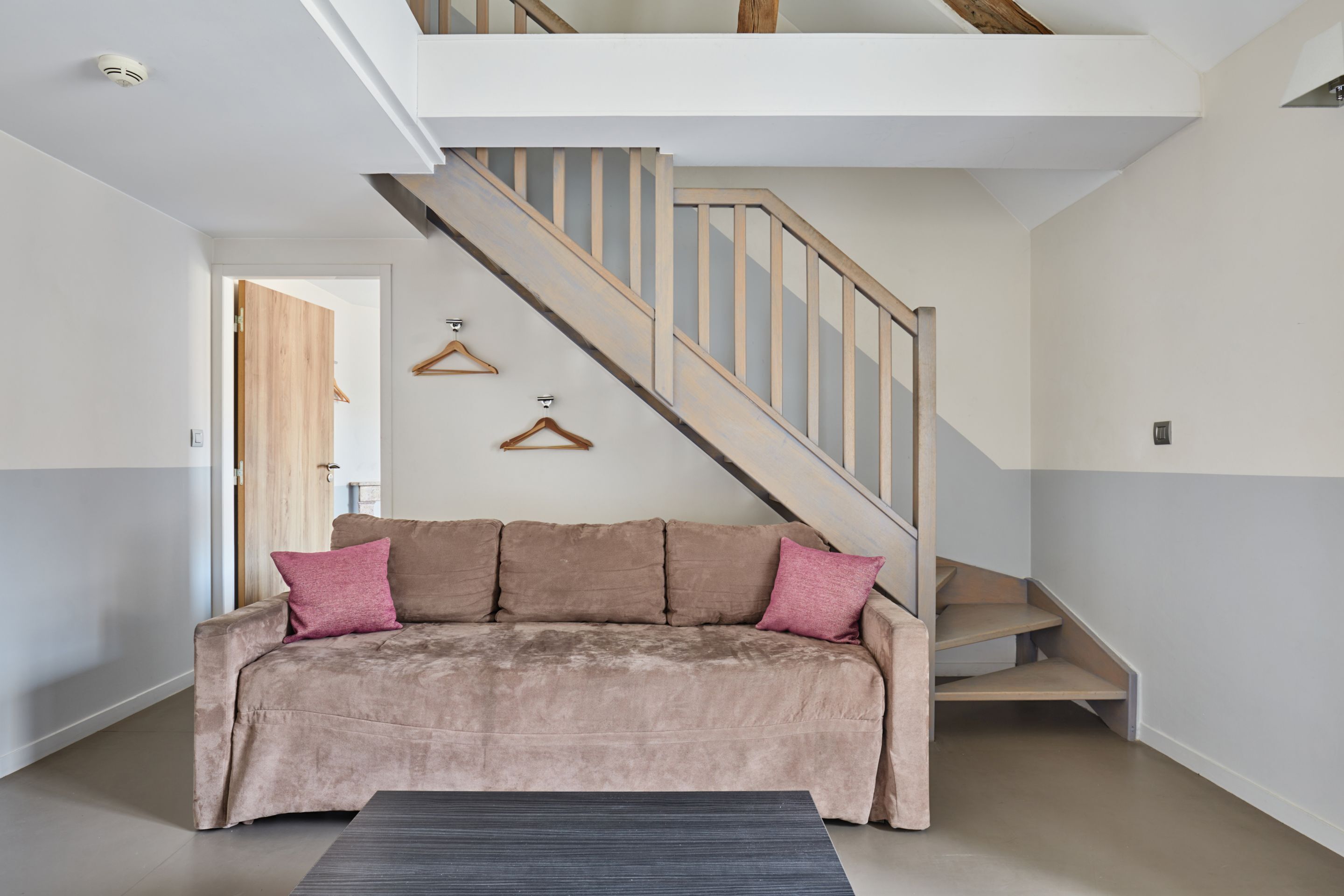 Una sala de estar moderna con un sofá color beige, una escalera de madera y una puerta que da a otra habitación.