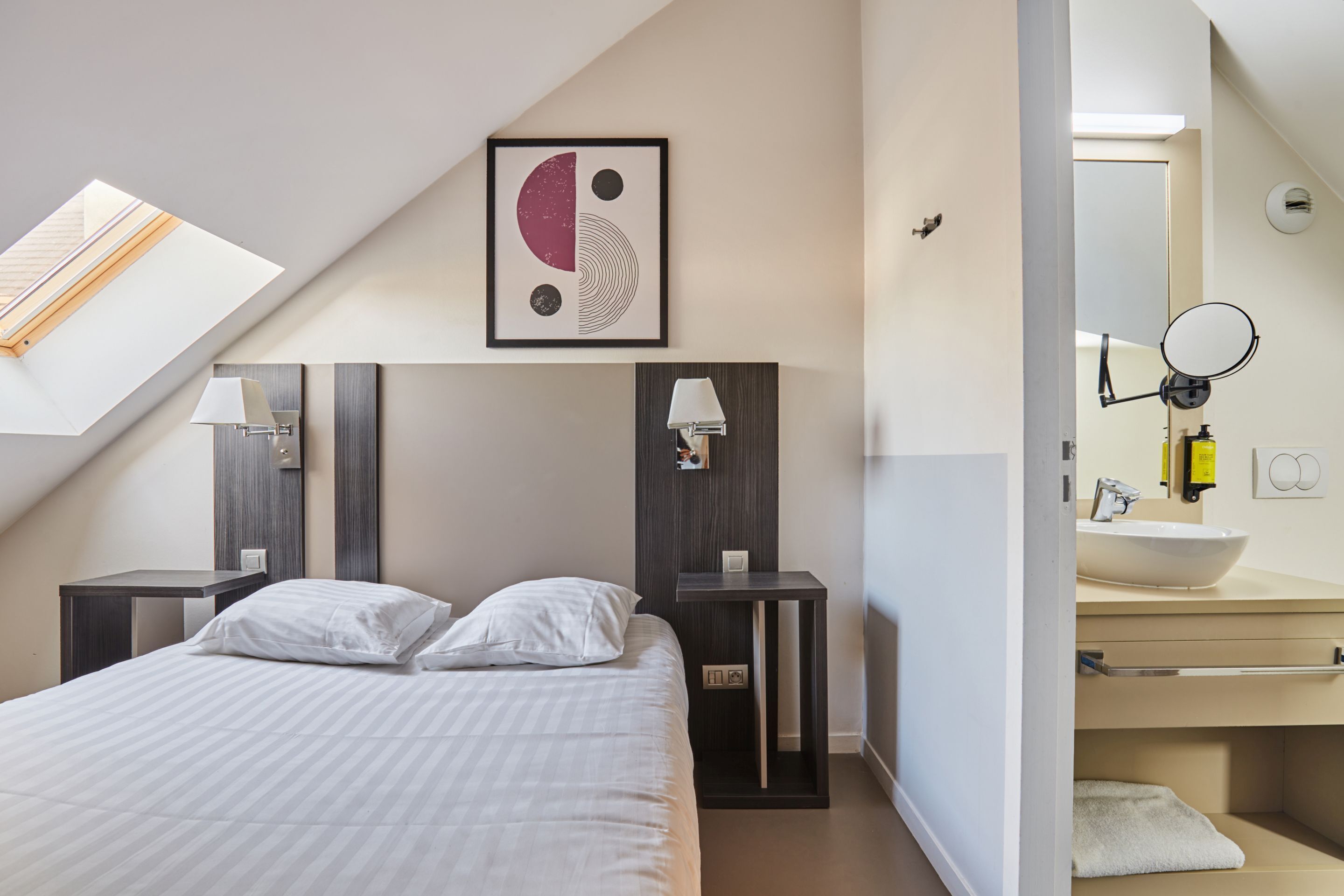 A modern hotel room with a skylight, a bed with white bedding, two bedside tables with lamps, a framed artwork on the wall, and a bathroom with a sink and mirror.