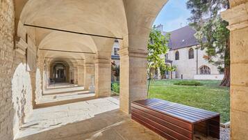 A stone corridor with arches, a bench, and a building with a lawn in the background.
