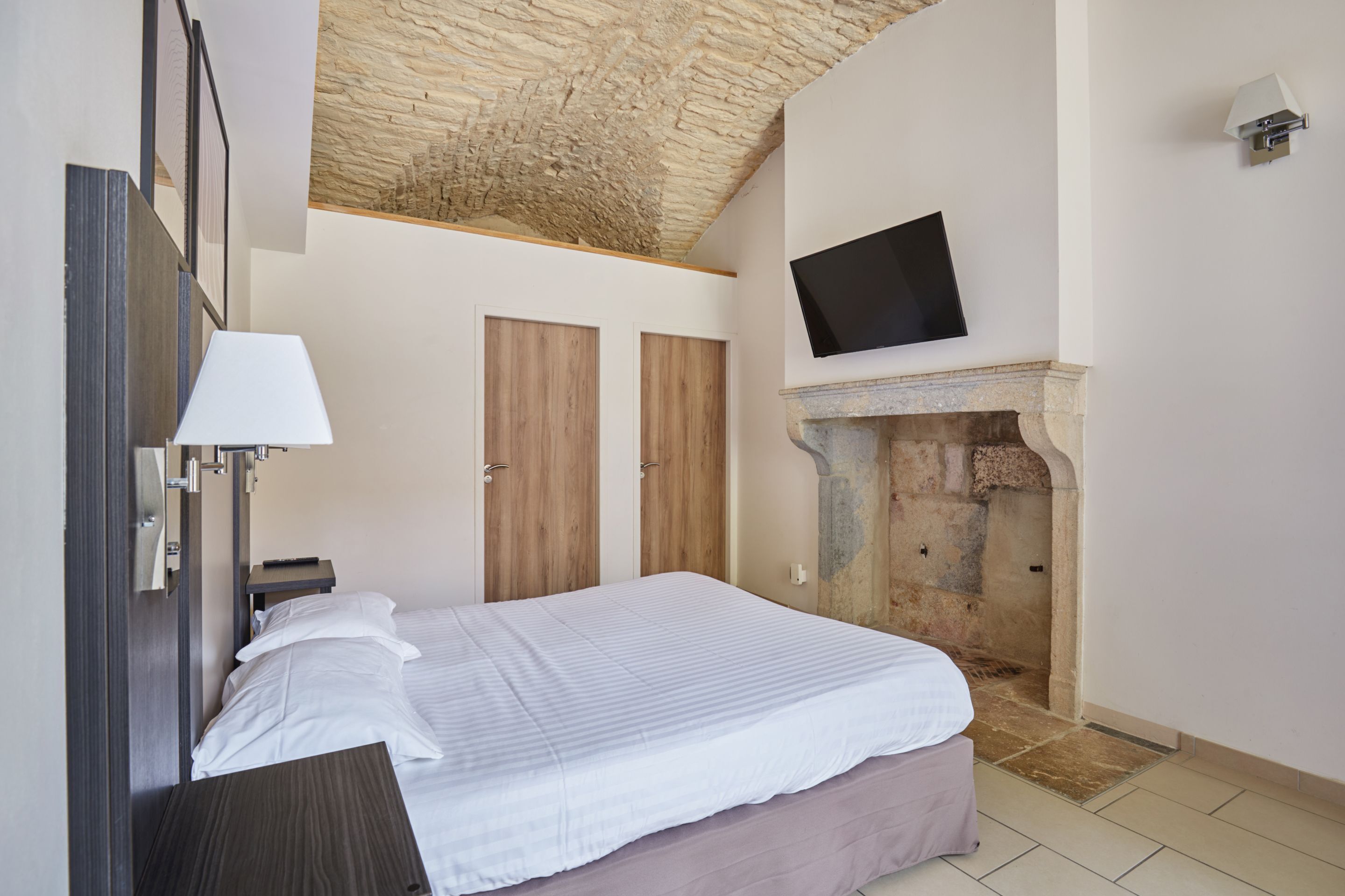 A bedroom with a bed, a mounted TV, a stone fireplace, and wooden doors.