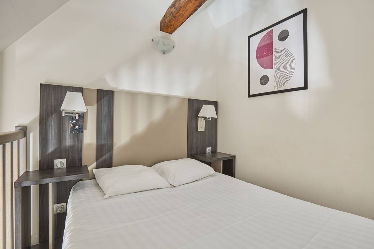 Quarto com cama, duas mesas de cabeceira com abajures, cabeceira de madeira e uma obra de arte abstrata na parede.