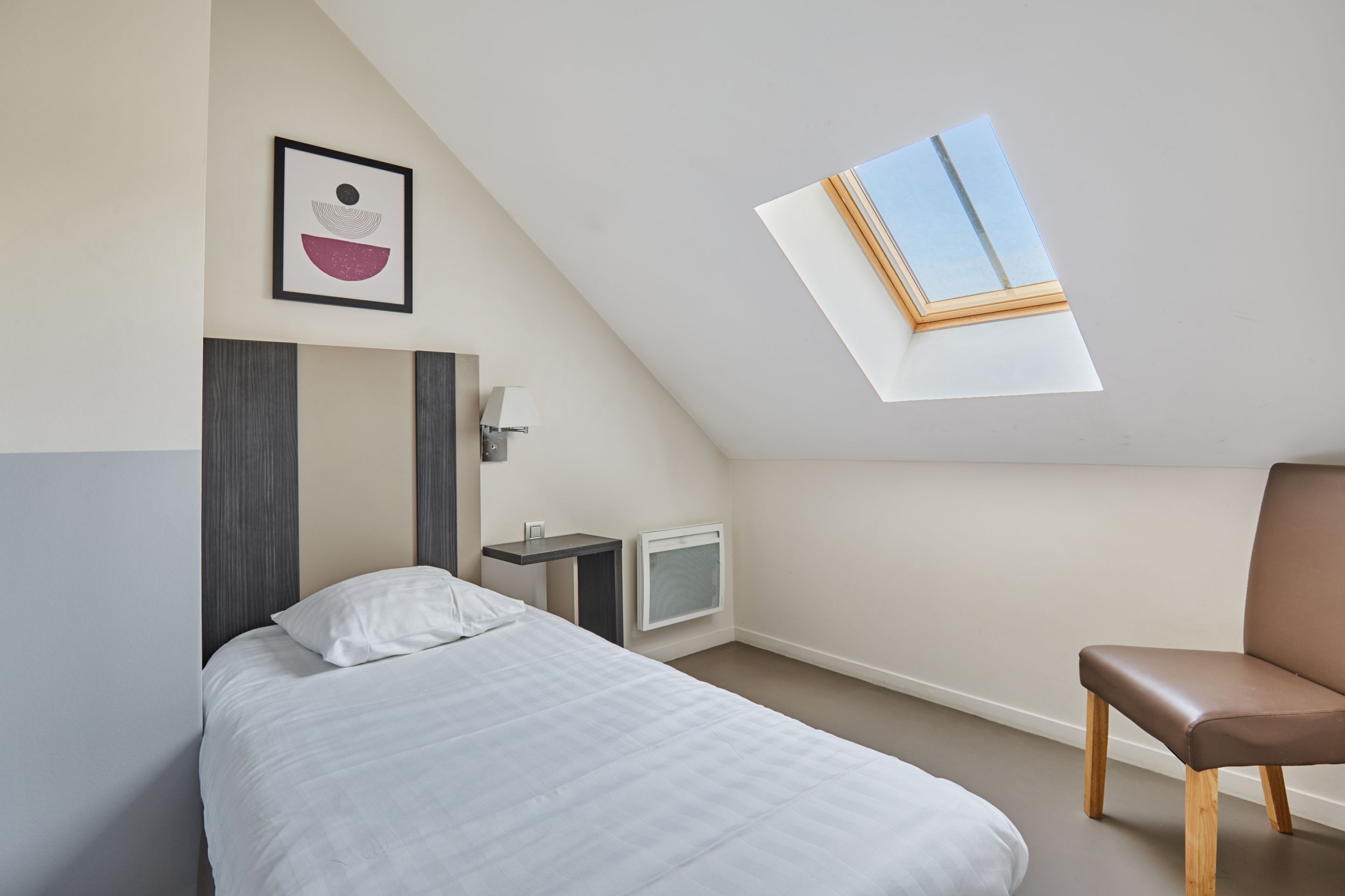 Un dormitorio con cama, una silla, mesita de noche, lámpara, televisor y tragaluz.