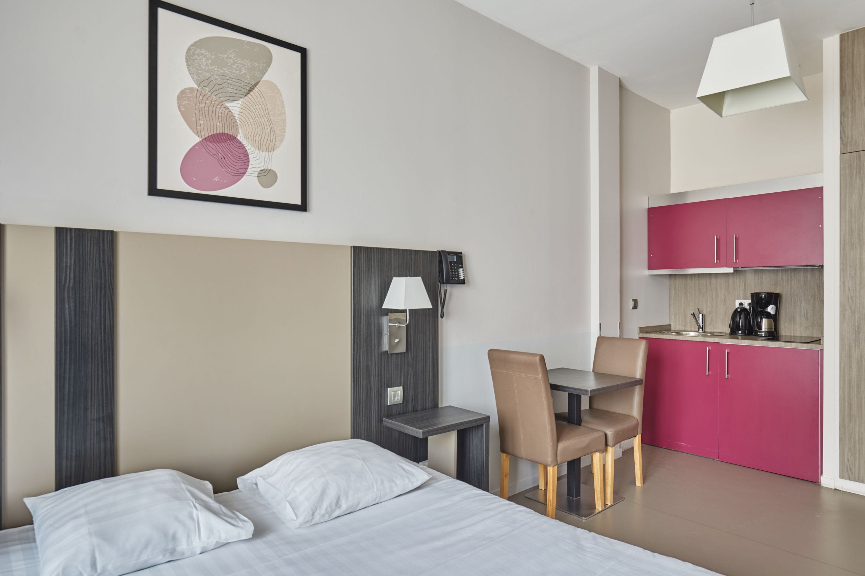 A hotel room with a bed, a desk, a chair, a small table, a lamp, a telephone, and a kitchenette with pink cabinets.