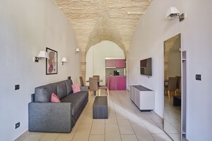 Uma sala de estar moderna com um sofá cinzento, uma mesa de centro e uma área de jantar com cadeiras e uma mesa. Uma cozinha aparece ao fundo, com armários cor-de-rosa e uma TV fixada na parede.