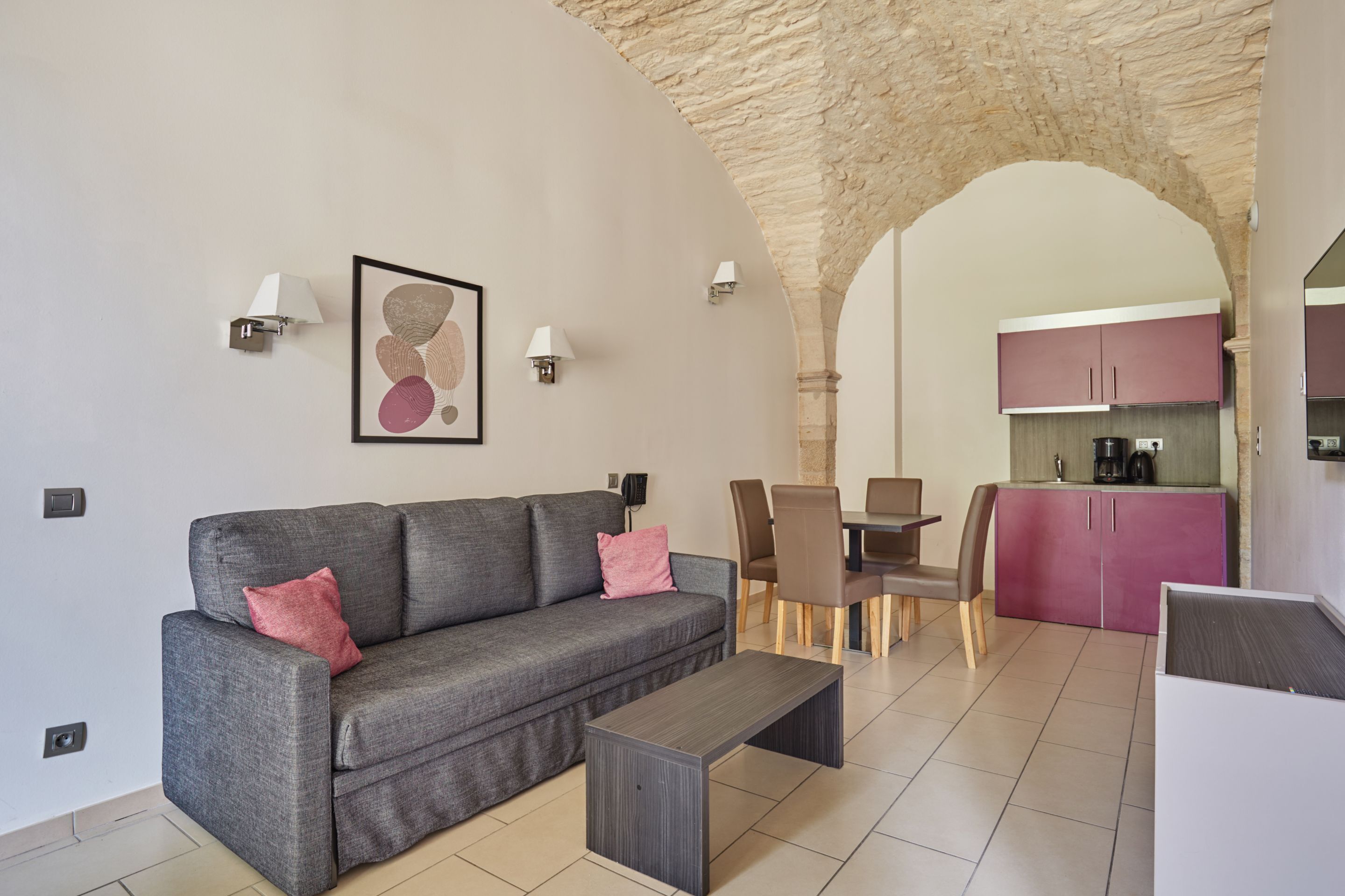 A modern living room with a grey sofa, a coffee table, and a dining area with chairs and a table. There is a kitchen with pink cabinets in the background.