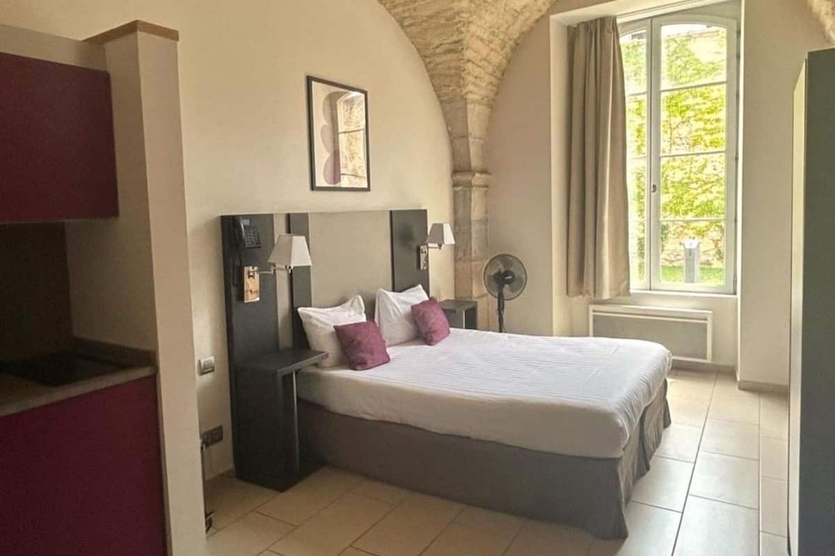 Quarto com cama, mesas de cabeceira, abajur, ventilador e janela com cortinas.