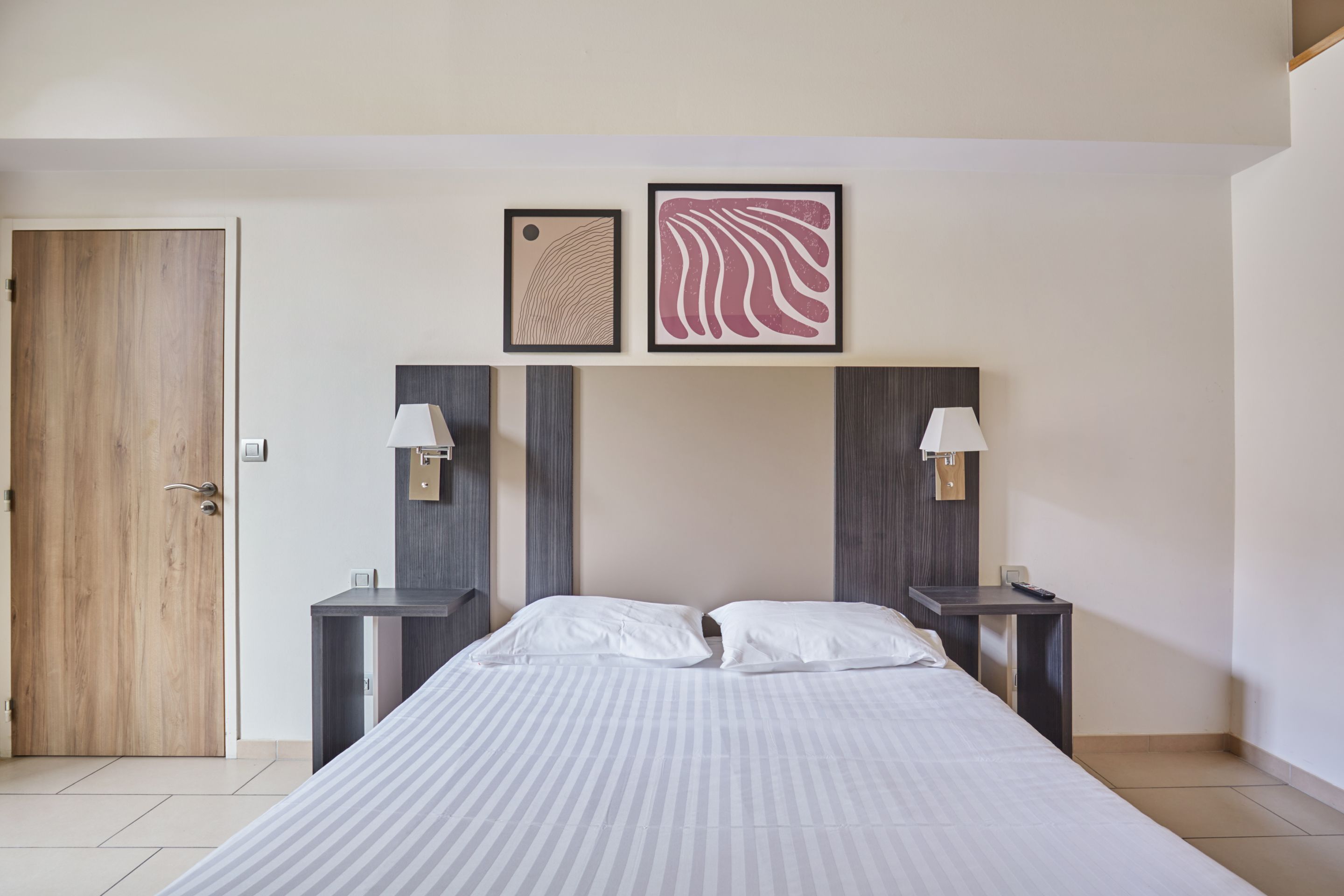 Une chambre à coucher bien rangée, avec un grand lit, deux tables de chevet avec des lampes, une porte en bois et deux œuvres d’art encadrées accrochées au mur.