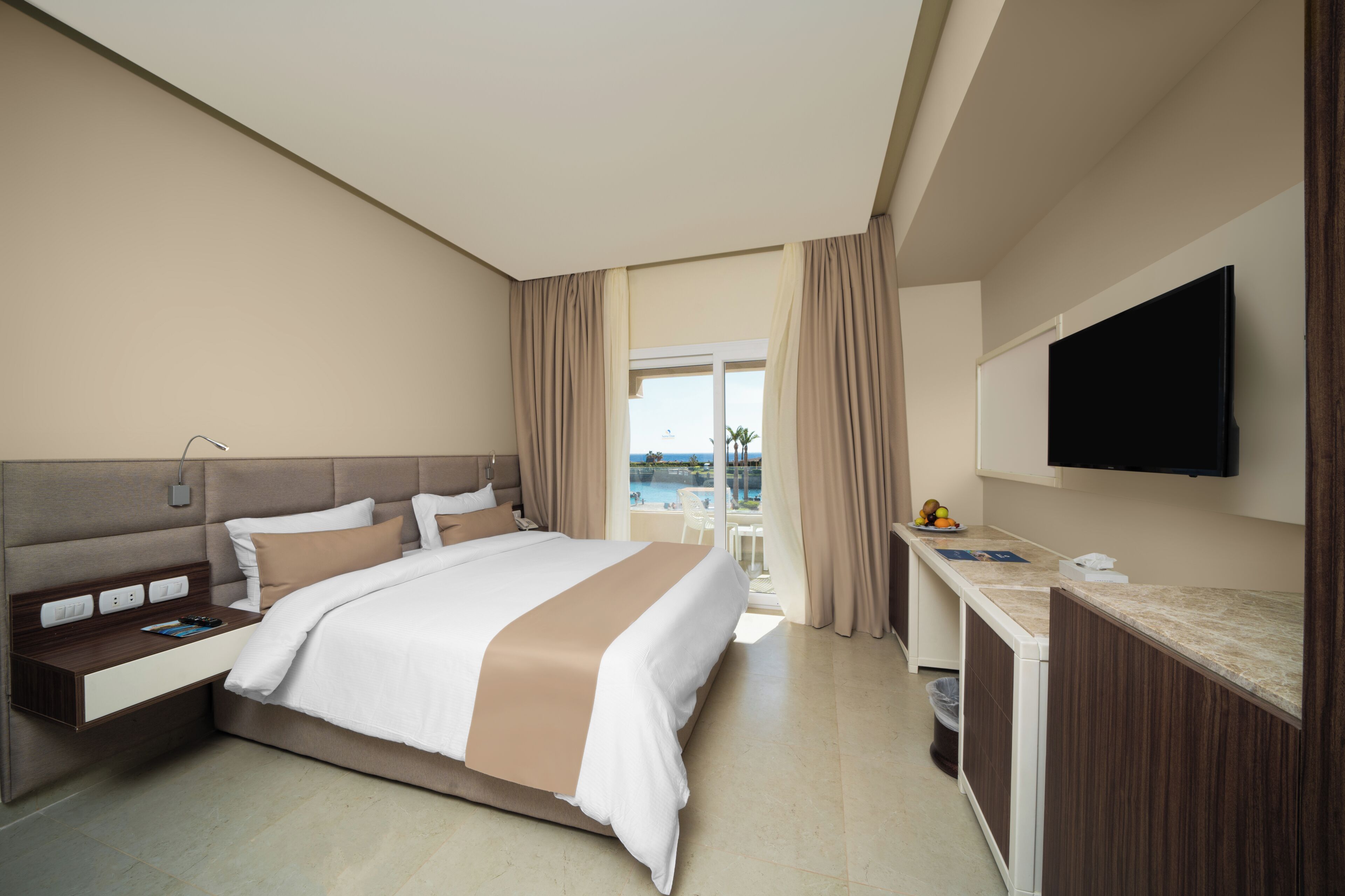 A hotel room with a bed, bedside table, TV, and a view of a pool.
