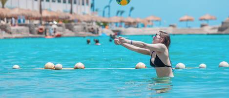 Una persona jugando al voleibol en una piscina azul cristalina con boyas, una playa y edificios al fondo.