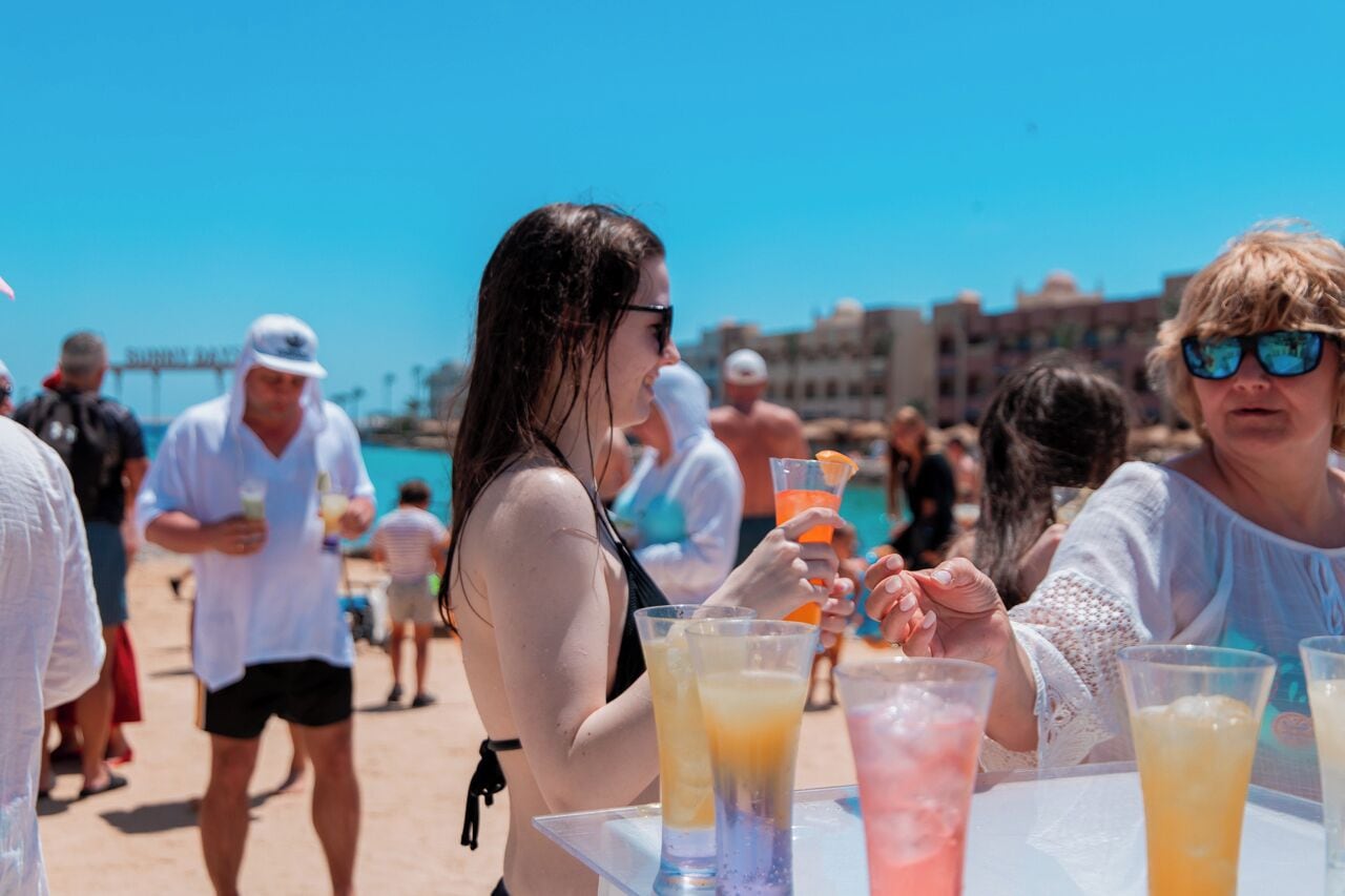 Un bar en la playa con mesas, bebidas y gente vestida informalmente, bajo un cielo azul despejado.