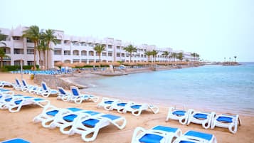 A beach with rows of sun loungers, a pool, and a hotel building with balconies.