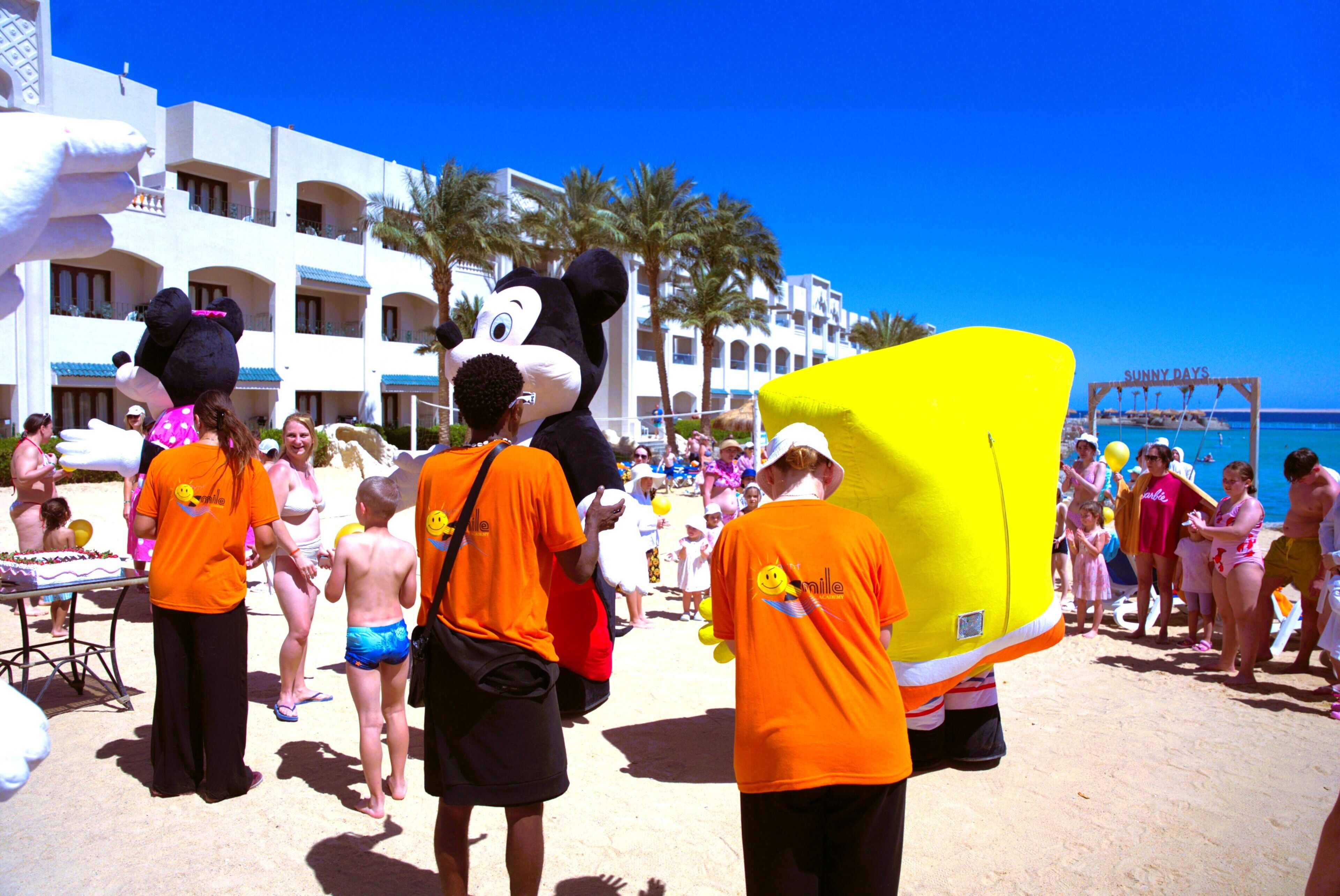 Une scène de plage avec des personnes, une grande structure jaune et un bâtiment doté de balcons en arrière-plan.