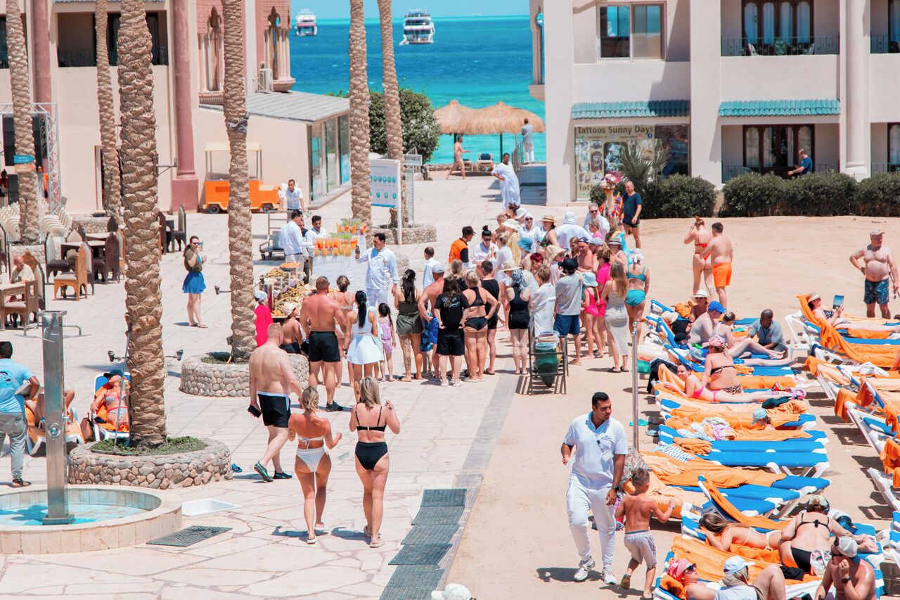 A crowded beachside area with lounge chairs, palm trees, and a pool.