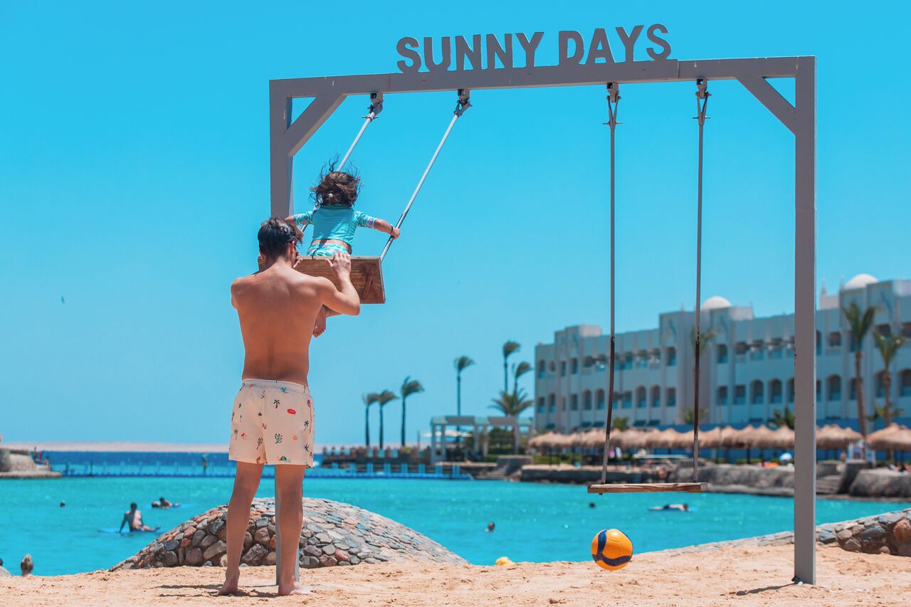 Cena de praia com baloiço identificado como 'SUNNY DAYS', piscina e um edifício ao fundo.