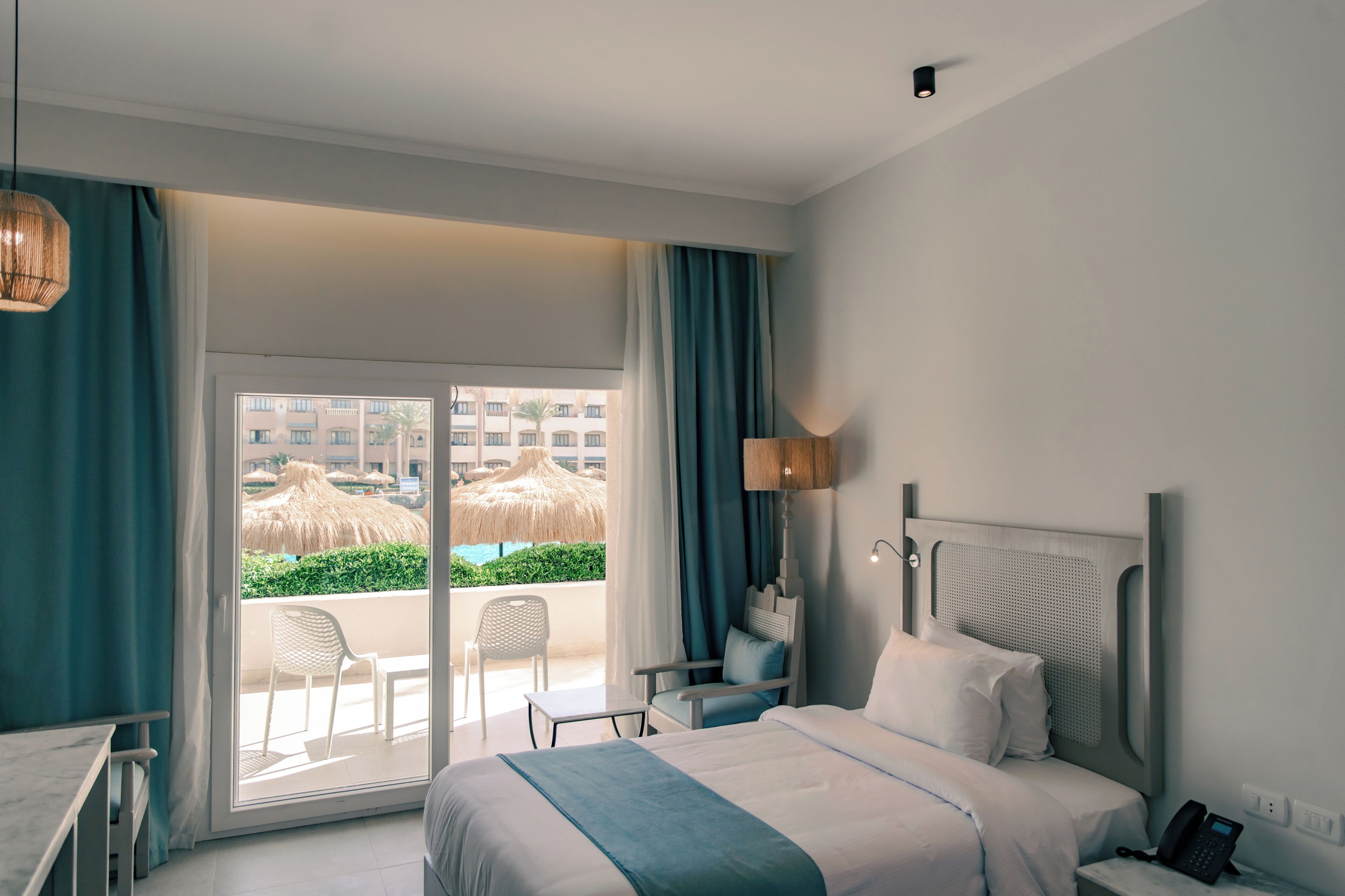 A hotel room with a bed, a desk, a chair, a phone, and a view of a pool and buildings.