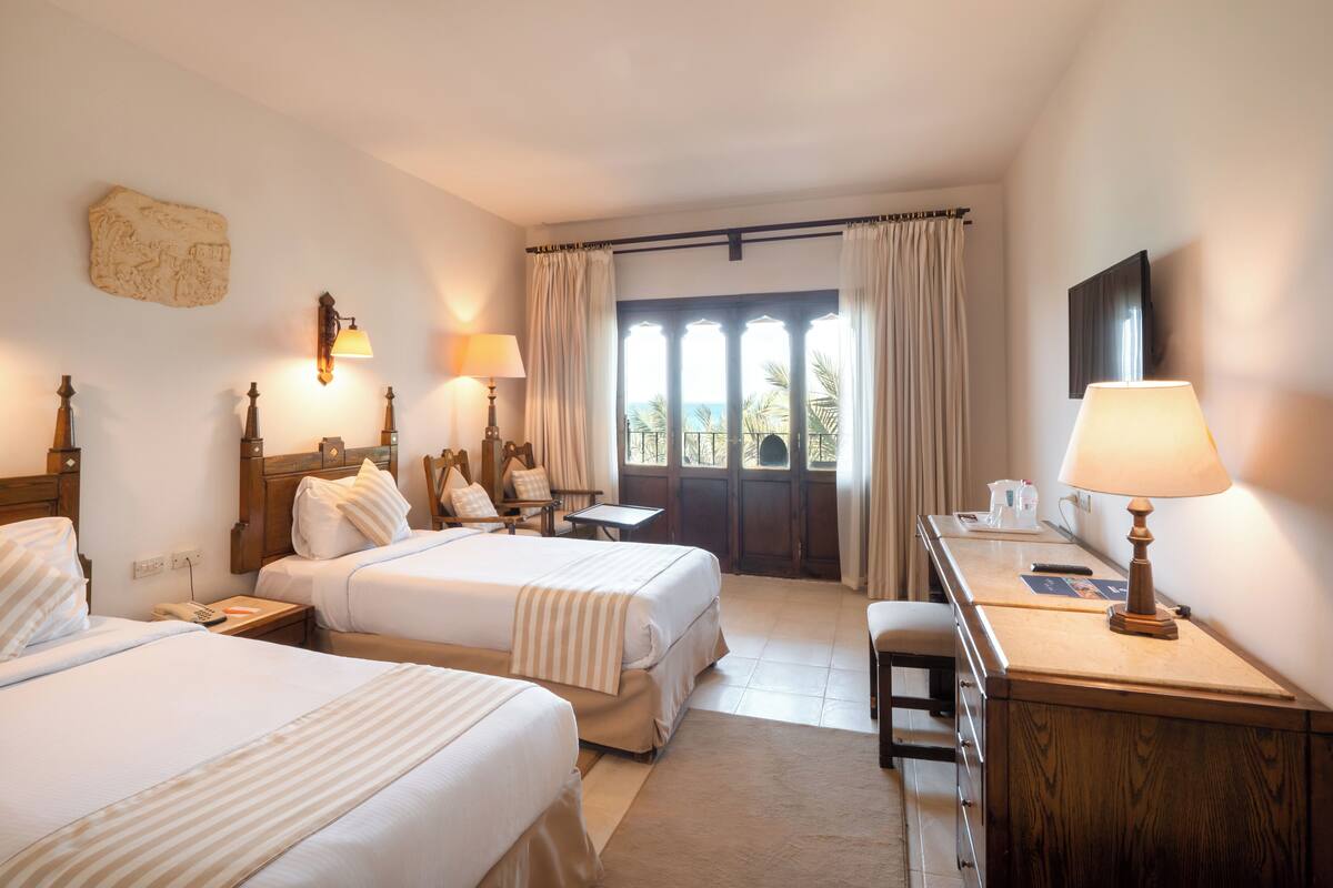 Une chambre d’hôtel avec deux lits, un bureau avec une lampe, une chaise et une vue sur la mer par la fenêtre.