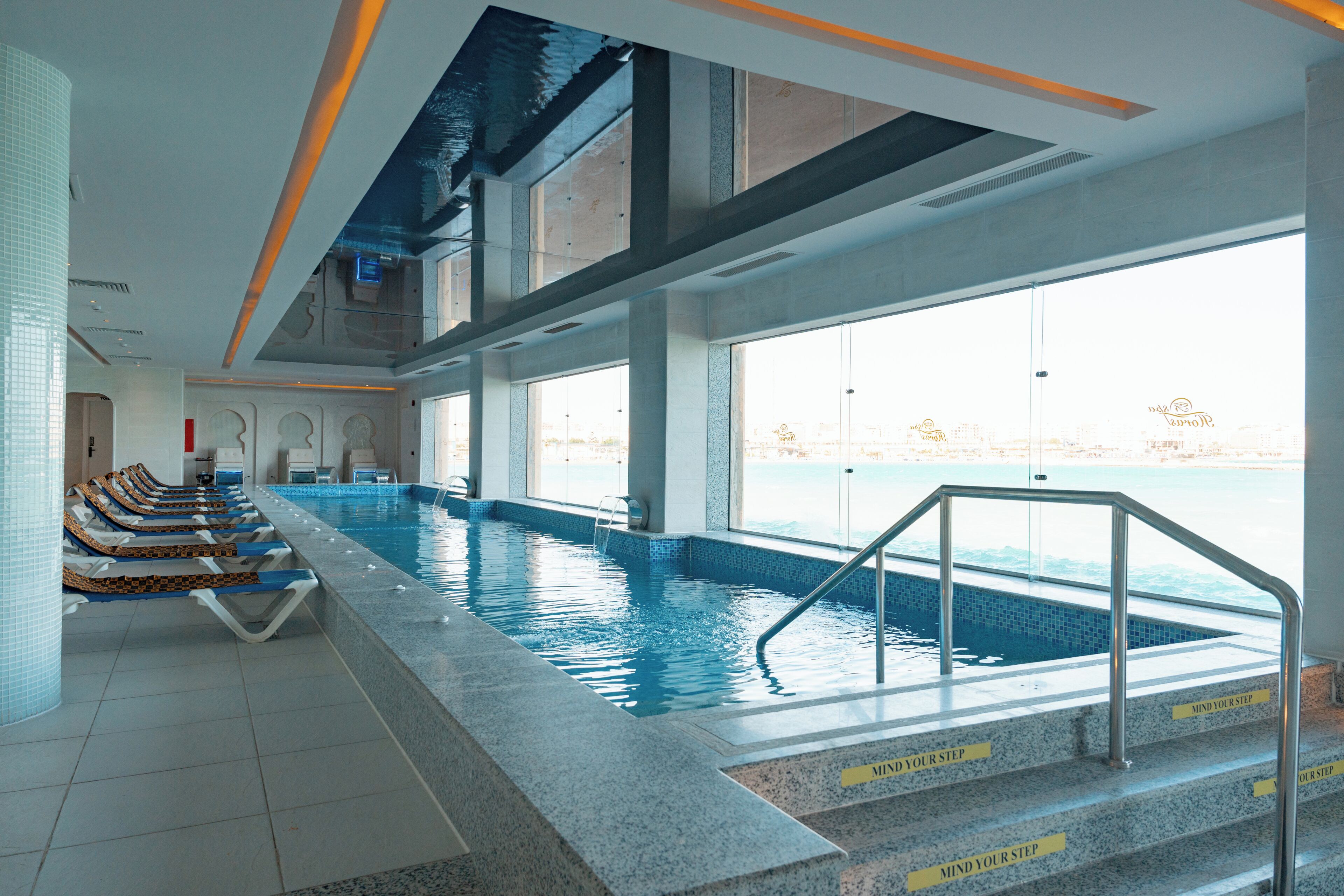 Une piscine intérieure avec des parois en verre transparent, un sol carrelé et des marches menant à l’intérieur de la piscine.