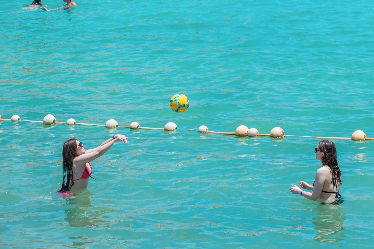 Une activité aquatique où les participants, vêtus de maillots de bain, doivent franchir une barrière flottante à l’aide d’un ballon.