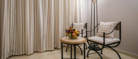Una habitación con una cortina a rayas, una mesa redonda con un frutero y una silla con un cojín de cuadros.