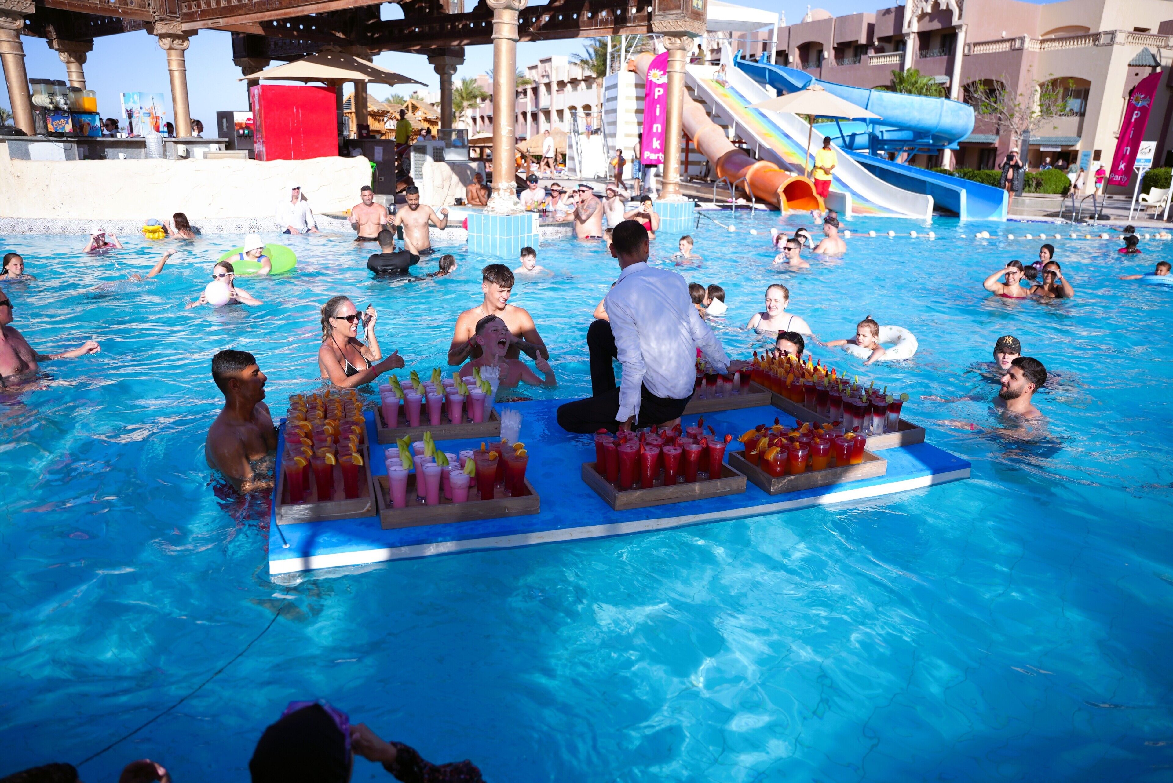 Zona de piscina con gente nadando y relajándose, tobogán acuático y zona de bar con bebidas.