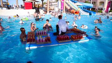 Zona de piscina con gente nadando y relajándose, tobogán acuático y zona de bar con bebidas.