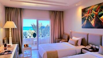 A hotel room with two beds, a desk with a lamp, a painting, and a view of the ocean.