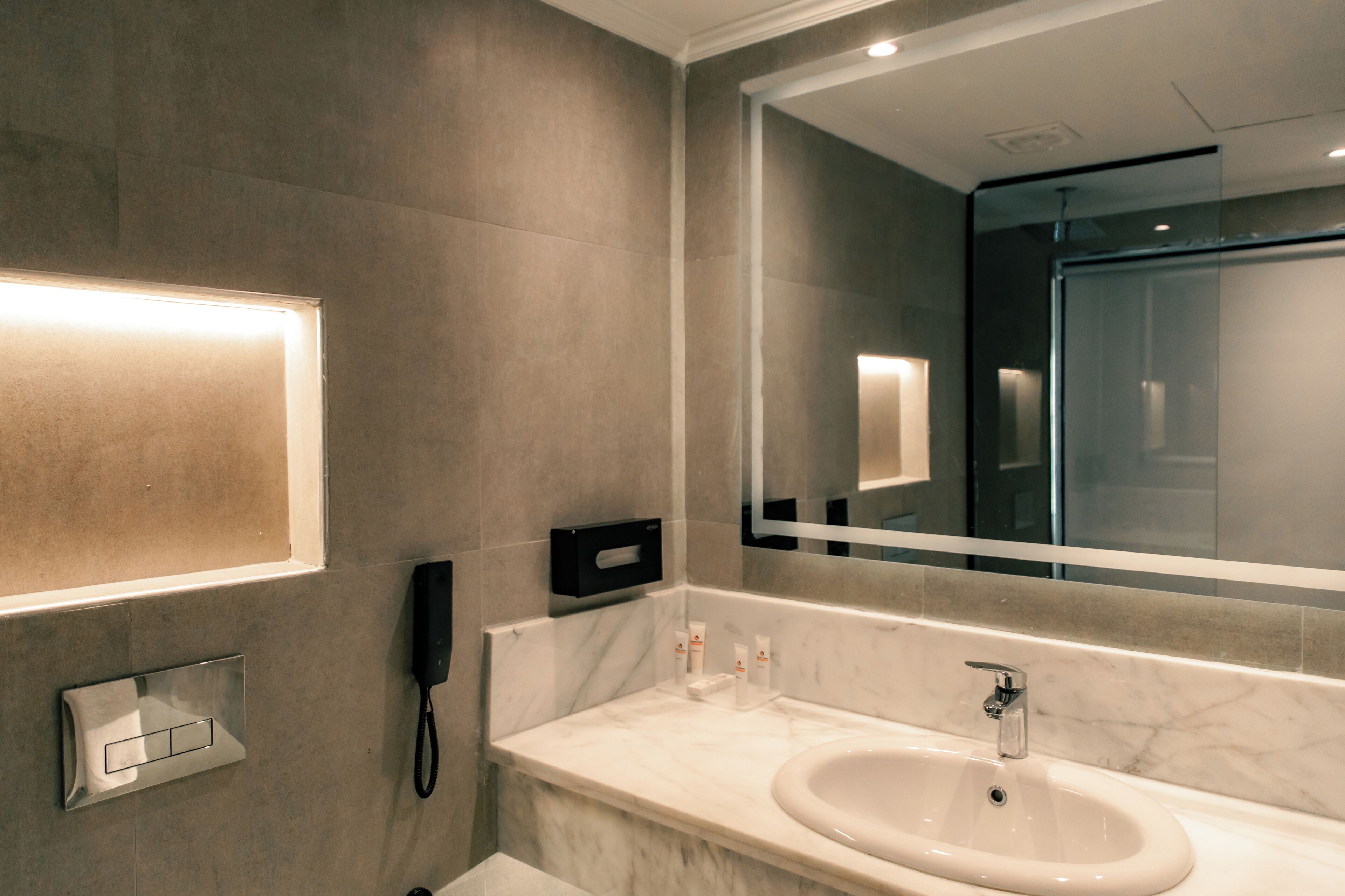 A modern bathroom with a large mirror, a sink, a telephone, and a lighted wall panel.