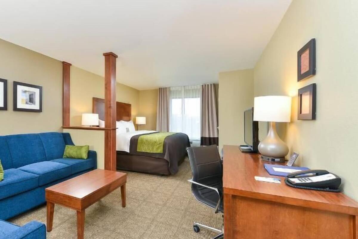 A hotel room with a blue sofa, a wooden coffee table, a desk with a lamp, a bed with a green blanket, and two framed pictures on the wall.