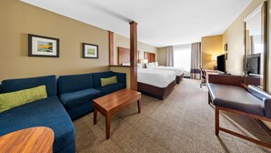A hotel room with a blue sofa, a wooden coffee table, a bed with white linens, a TV, and a large mirror. - Comfort Inn & Suites West - Medical Center (Rochester)